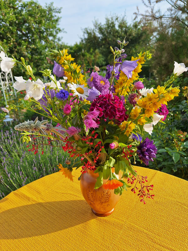 Vielfltige Gartenblumen in krftigen Farben wie Gelb, Pink, Violett und Blau in einem dichten Pflanzenstrau. Der Strau steht in einer terrakottafarbenen Vase auf einem Gartentisch mit gelber Tischdecke inmitten eines naturnahen Kleingartens